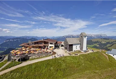 Eine malerische Berglandschaft mit einer Hüttenanschrift und einer kleinen Kirche. Viele Menschen genießen die Aussicht auf der Terrasse.