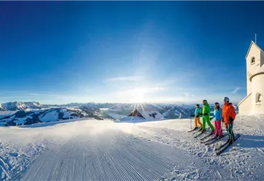 A winter mountain landscape with skiers in front of a small white chapel. The sky is clear and bright blue, while the sun shines on the horizon.