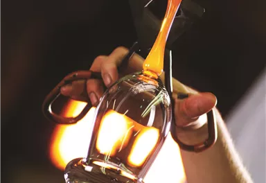 Ein Künstler formt ein Glasobjekt aus glühendem Glas. Im Hintergrund ist eine warme, diffusive Beleuchtung zu sehen.