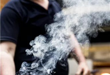 Ein Glasbläser arbeitet konzentriert mit heißem Glas. Rauch steigt auf, während er die orangefarbene Glasmasse formt.