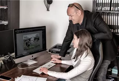 A man stands behind a woman and looks at a computer screen. Both appear to be working on a project.