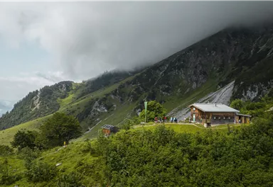 A charming mountain cabin is situated on a green meadow, surrounded by majestic mountains and dense clouds. The scene radiates tranquility and closeness to nature.