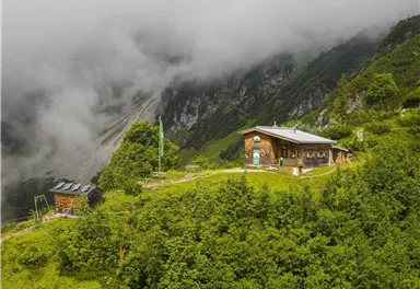 A rustic cabin surrounded by green meadows and mountains. The sky is cloudy and the landscape appears calm and idyllic.