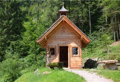 A small wooden chapel in the woods, surrounded by green trees. A narrow path leads to the door of the chapel.