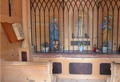 A small chapel with wooden cladding and enclosed figures. In the background, there are religious statues and a small floral decoration.