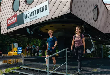 Zwei Wanderer gehen eine Treppe hinunter von einer modernen Bergbahnstation. Im Hintergrund sind weitere Menschen und eine malerische Landschaft zu sehen.