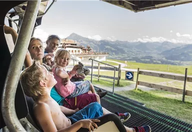 Eine Gruppe von Kindern und Erwachsenen sitzt in einer Seilbahn und genießt die Aussicht auf die Berge. Im Hintergrund sind grüne Wiesen und eine alpine Landschaft zu sehen.