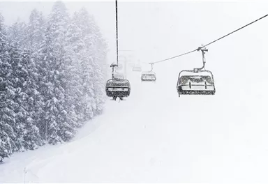 Eine schneebedeckte Landschaft mit einem Skilift in der Mitte. Die Bäume im Hintergrund sind von Schnee umgeben und die Sicht ist eingeschränkt.