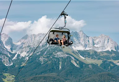 Zwei Personen sitzen in einer Gondel auf dem Weg über den Bergen. Die Landschaft besteht aus grünen Wäldern und majestätischen Gipfeln unter einem klaren Himmel.