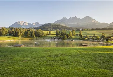 A picturesque landscape with a tranquil lake and gentle hills. In the background, majestic mountains rise under a clear sky.
