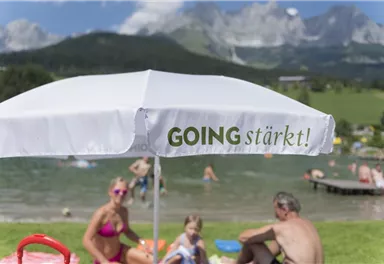 A beautiful day at a lake with people relaxing in the water. A beach umbrella with the inscription "GOING strengthens!" is in the foreground.