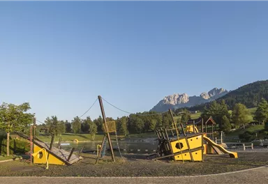 A playground with a three-dimensional climbing structure in the shape of a ship. In the background, there are mountains and a clear blue sky.