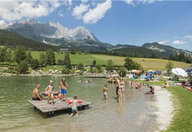 A beautiful lake with many people playing in the water and standing on a dock. In the background, green meadows and mountains can be seen.