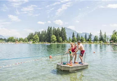 A cheerful day by the water with children on a wooden jetty. In the background, you can see green trees and mountains under a clear sky.