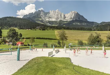 A beach volleyball court in a picturesque landscape with mountains in the background. In the foreground, people are playing volleyball under a blue sky.