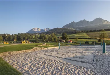 A sand court outdoors, surrounded by mountains and forests. The sky is clear and the landscape is green.