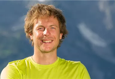 Ein lächelnder Mann trägt ein leuchtend grünes Shirt. Im Hintergrund sind verschwommene Berge zu sehen.