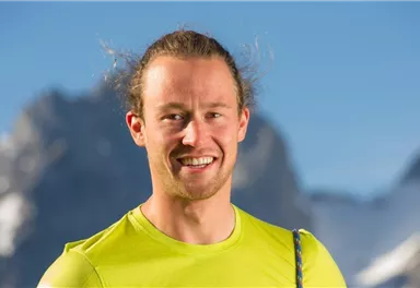 A smiling man in a brightly colored yellow shirt. In the background, mountains with snow-capped peaks can be seen.