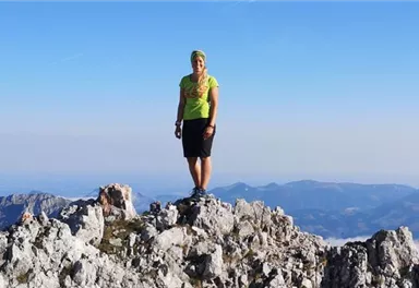 A person stands on a rocky summit with a magnificent view of the mountains and the sky. They are wearing a bright green T-shirt and sporty shorts.