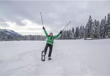 A cheerful person stands in the snow and raises a ski pole in the air. In the background, snow-covered trees and a wintry landscape can be seen.