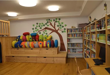 Ein gemütlicher Lese- und Spielbereich mit bunten Kissen und einer Wanddekoration eines Baumes. Regale mit Büchern sind ebenfalls vorhanden.