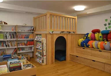 Ein einladender Raum mit einem Lesebereich, Bücherregalen und einem gemütlichen Rückzugsort. Bunte Kissen bieten Platz zum Sitzen und Entspannen.