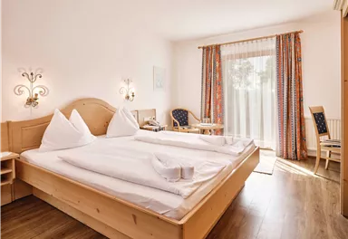 Ein gemütliches Schlafzimmer mit einem großen Bett und hellen Kopfkissen. Das Zimmer hat ein Fenster mit Vorhängen und einen kleinen Tisch mit Stühlen.