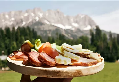 Ein Holzbrett mit verschiedenem Aufschnitt, Käse und frischem Gemüse vor einer beeindruckenden Bergkulisse. Die Szene strahlt eine gemütliche, alpine Atmosphäre aus.