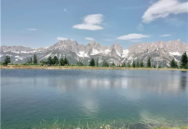 A tranquil mountain lake with smooth water and majestic mountains in the background. The sky is clear with a few clouds.