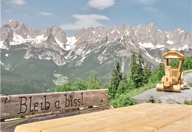 A beautiful mountain landscape with snow-covered peaks. In the foreground stands a wooden sign with the inscription "Stay a little" and a small wooden vehicle.