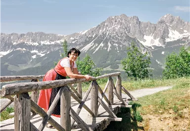 A woman in traditional attire stands on a wooden bridge in the mountains. In the background, snow-capped peaks and green trees can be seen.