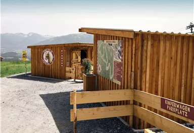 A wooden building at the parking lot with an information wall about the surroundings. In the background, mountains and a clear sky can be seen.