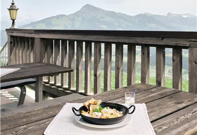 A delicious plate of food is placed on a rustic table on the terrace. In the background, green mountains and a clear sky are visible.