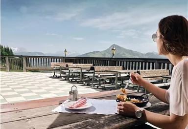 A woman sits at a table enjoying her meal with a view of the mountains. The sky is clear and the terrace is well-furnished.