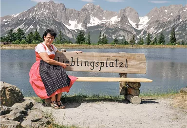 A woman is sitting on a bench with the inscription "Favorite Place" by the water. Impressive mountains and a clear sky can be seen in the background.