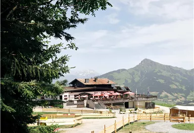 Ein rustikales Gebäude in den Bergen mit einer Terrasse, auf der Gäste sitzen. Im Hintergrund sind grüne Hügel und ein klarer Himmel zu sehen.