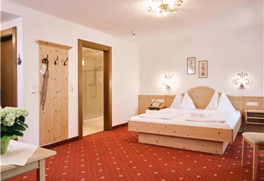 Ein helles Zimmer mit zwei Betten und einem roten Teppich. Es gibt Holzdetails und einen kleinen Tisch mit einer Blume.