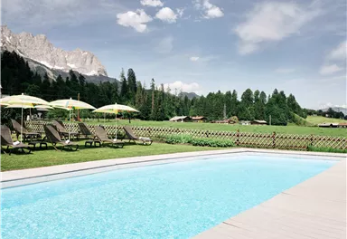 Ein schöner Pool mit klarem Wasser, umgeben von grünen Wiesen und Bergen. Liegen und Sonnenschirme laden zum Entspannen ein.