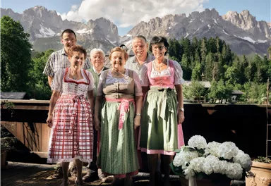 Eine Gruppe von sechs Personen in traditionellen Trachten steht vor einer beeindruckenden Berglandschaft. Im Hintergrund sind grüne Wälder und hohe Berge sichtbar.