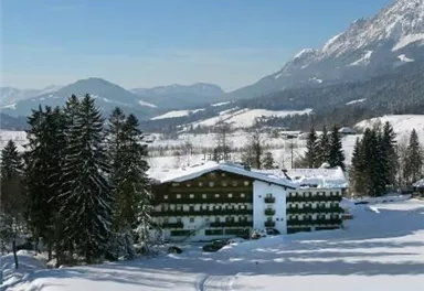 Ein malerisches Hotel in einer Winterlandschaft, umgeben von schneebedeckten Bäumen. Im Hintergrund sind die Berge unter einem klaren blauen Himmel zu sehen.