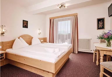 Ein gemütliches Hotelzimmer mit einem Doppelbett und Holzmöbeln. Die Fenster sind mit Vorhängen versehen und eine kleine Blumenvase steht auf dem Tisch.