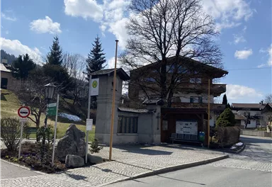 Ein charmantes Gebäude an einer ruhigen Straße, umgeben von Bäumen und einer schönen Landschaft. Der Himmel ist klar mit einigen Wolken.
