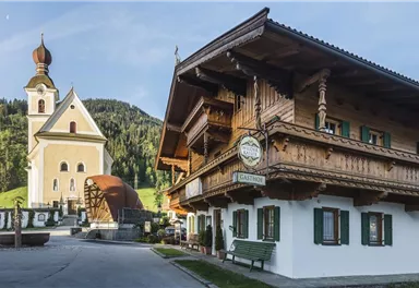 A traditional old inn with wooden decoration and green shutters stands next to a beautiful church. In the background, green hills and a clear sky are visible.