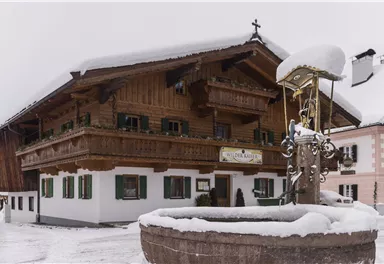Ein traditionelles Holzhaus im Schnee mit grünen Fensterläden. Im Vordergrund befindet sich ein Brunnen, umgeben von einer winterlichen Landschaft.