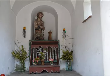 Eine kleine Kapelle mit einer Statue und einem Altar. Blumen und Kerzen schmücken den Raum.