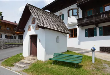 Ein kleines, weißes Gebäude mit einem schrägen Dach steht an der Straße. Neben dem Gebäude befindet sich eine grüne Bank.