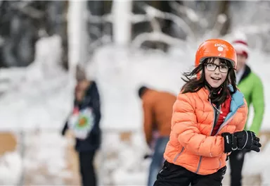 Ein fröhliches Mädchen in einem orangefarbenen Anorak fährt Schlittschuh auf einer gefrorenen Fläche. Im Hintergrund sind weitere Personen in winterlicher Kleidung zu sehen.