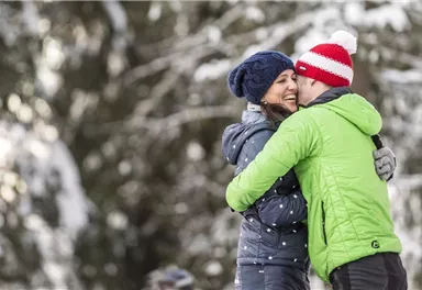Ein glückliches Paar umarmt sich in einer verschneiten Landschaft. Sie tragen warme Kleidung und lächeln einander zu.