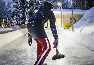 Eine Person steht auf einer Eisbahn und bereitet sich auf einen Wurf vor. Im Hintergrund sind verschneite Bäume und eine beleuchtete Hütte zu sehen.