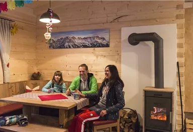 Eine gemütliche Stube mit einem Tisch, an dem drei Personen sitzen. Im Hintergrund gibt es einen Kamin und eine landschaftliche Wanddekoration.